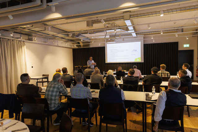 Innenaufnahme eines Macardo Brenn-Kurses: Mehrere Personen sitzen an Tischen in einem hellen Raum und blicken auf eine Leinwand mit Präsentation. Vor der Leinwand steht ein Mann und präsentiert den Kurs. Die Szene wirkt professionell.